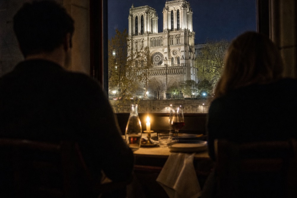 Dinner cruise passengers at a candlelit table with the lit facade of Notre-Dame visible through the window, no text, no logos, no people.