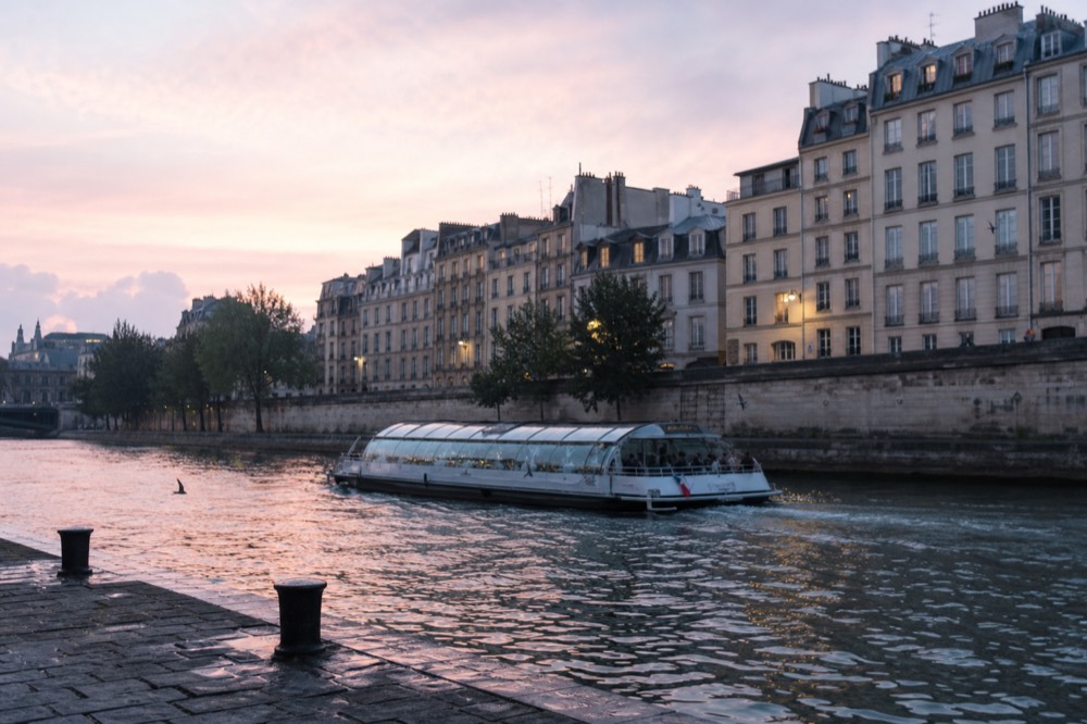 Glass-roofed dinner cruise boat on the Seine at dusk with Paris buildings behind, no text, no logos, no people