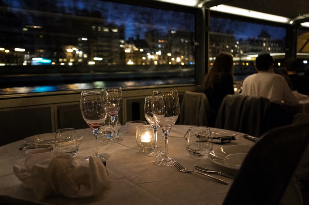 Set dinner table with wine glasses aboard a cruise boat, Paris lights blurred through windows, no text, no logos, no people