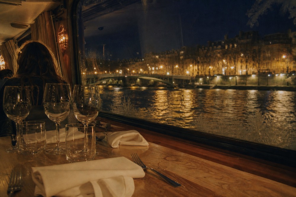 Table set for dinner aboard a boat with Paris lights beyond the window, no text, no logos, no people