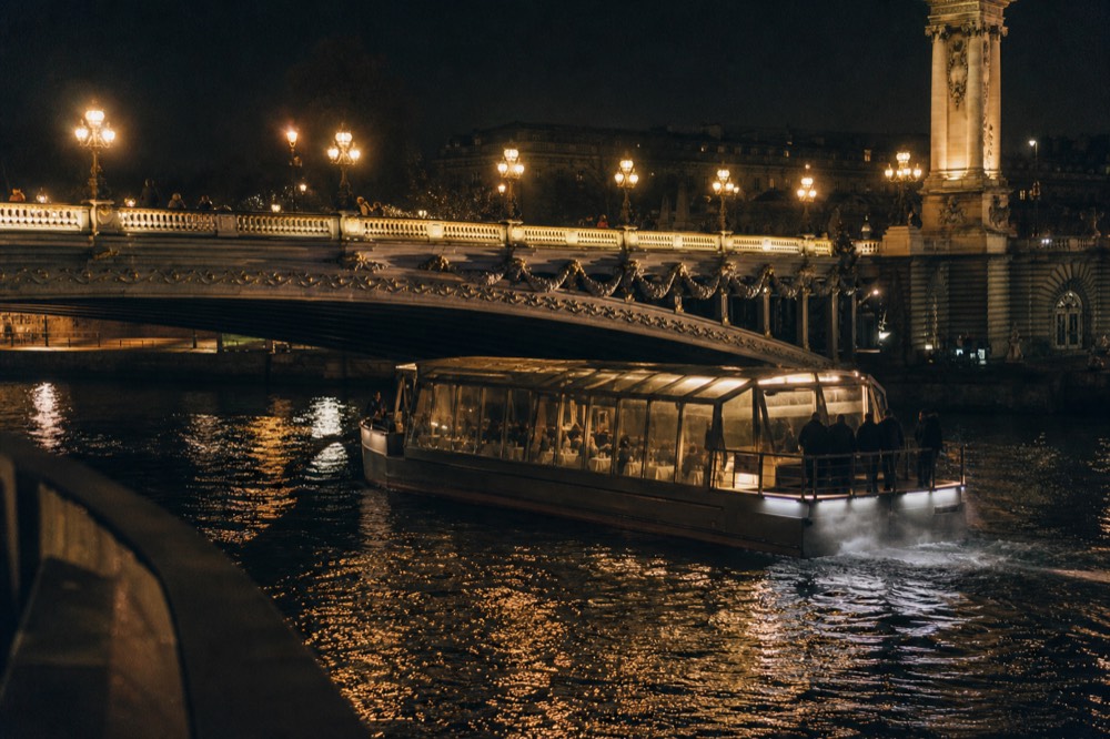 Dinner cruise boat passing under a Seine bridge at night, no text, no logos, no people