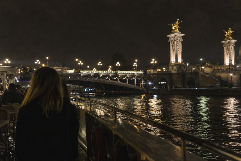 Dinner cruise boat lit up at night passing illuminated Parisian bridges, no text, no logos, no people