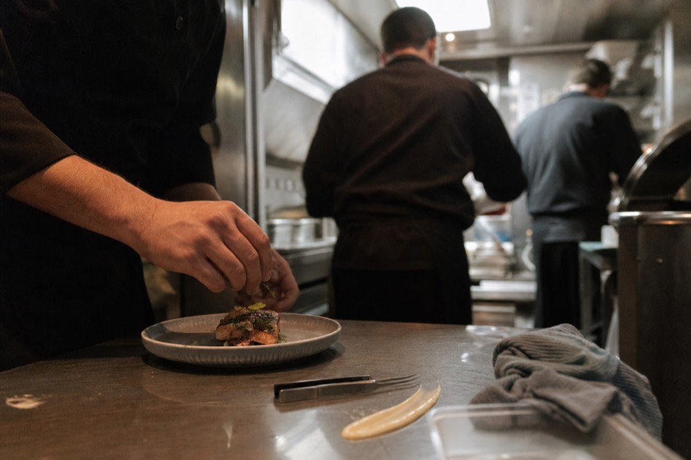 A chef plating an intricate fine-dining course in a compact galley kitchen, no text, no logos, no people