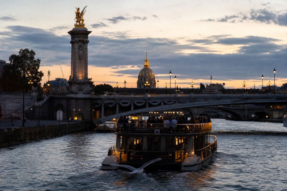 An illuminated Seine dinner cruise boat passing under a Paris bridge at dusk, no text, no logos, no people