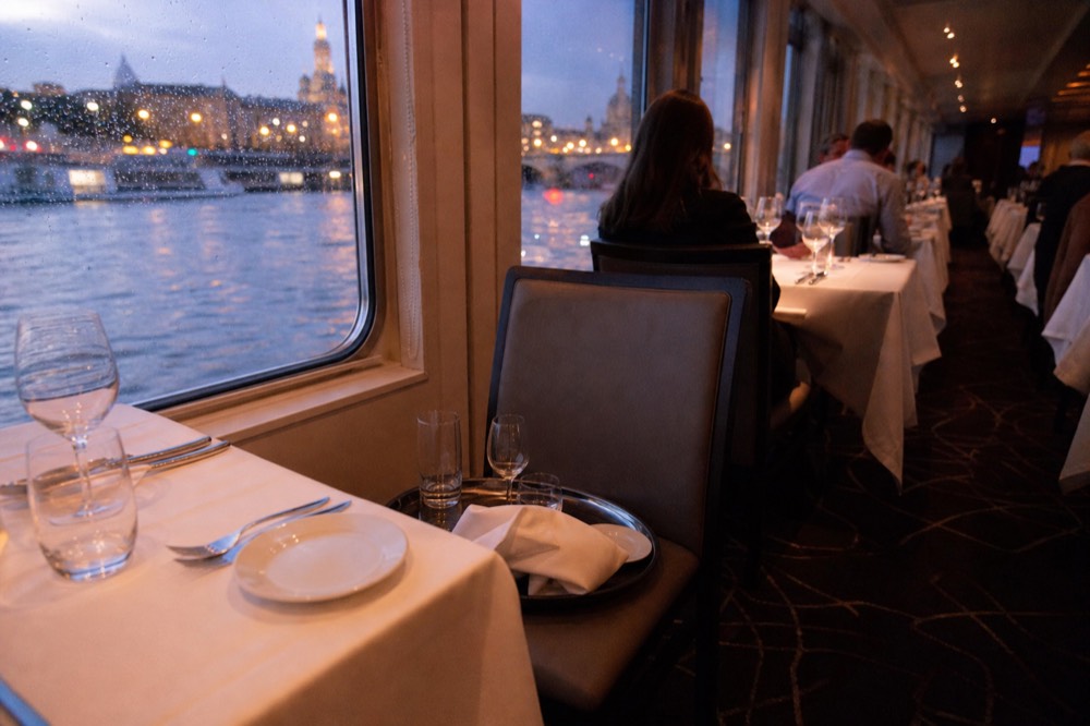 A riverboat dining room with set tables and city lights through the windows, no text, no logos, no people
