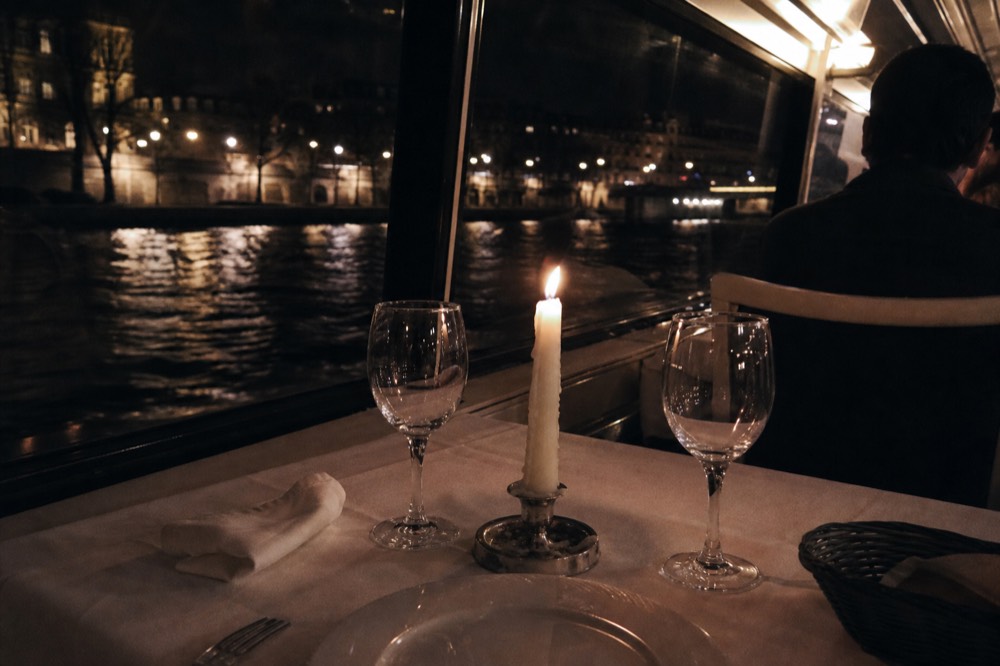 Candlelit dinner table set with wine glasses beside a window overlooking the Seine at night, no text, no logos, no people