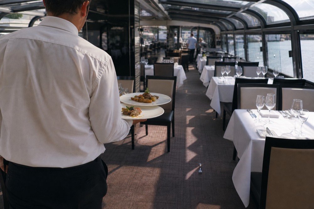 Waiter serving a plated three-course dinner inside an illuminated glass-covered cruise boat, no text, no logos, no people