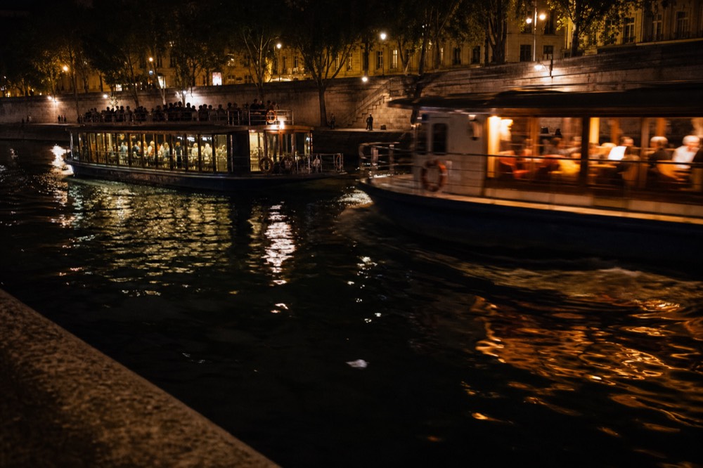 Two dinner cruise boats passing each other on the Seine at night, city lights reflecting on the water, no text, no logos, no people