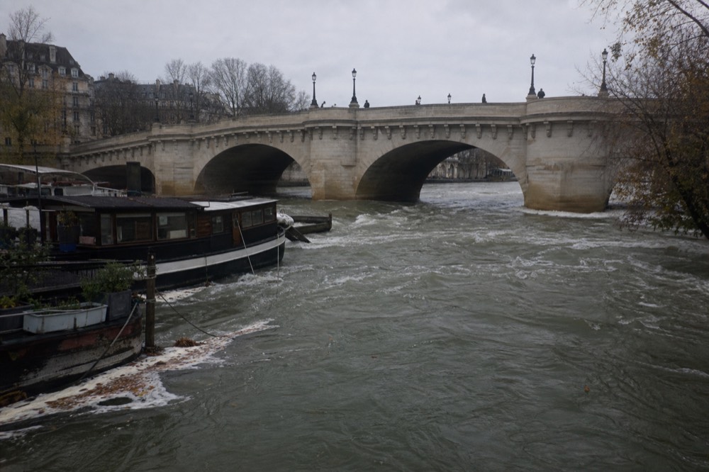 A high, fast-flowing Seine under a bridge with moored boats, overcast light, no text, no logos, no people
