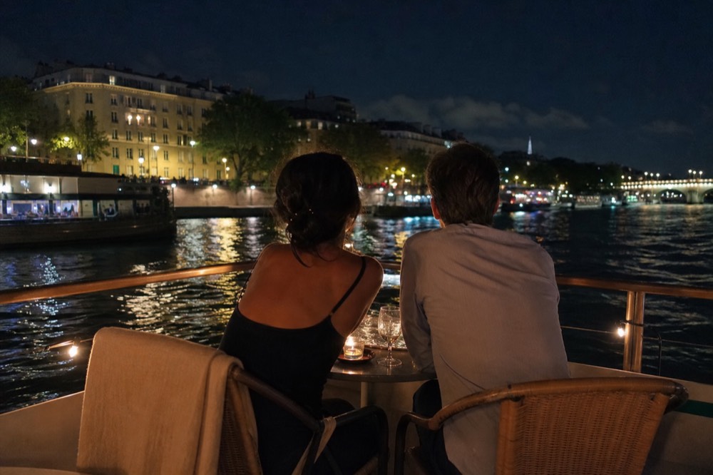 A couple at a candlelit table on a boat deck with Paris lights reflecting on the Seine, no text, no logos, no people