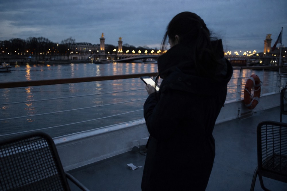 A passenger checking a phone on a dinner cruise deck at dusk in Paris, no text, no logos, no people