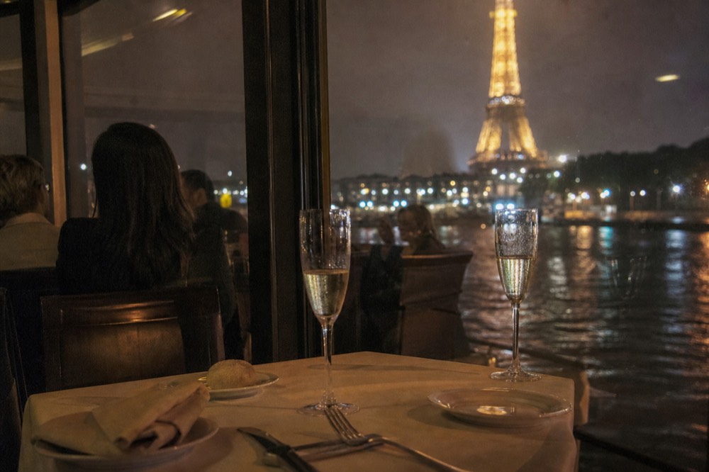 A dining table on a Seine dinner cruise with Champagne flutes and a view of the Eiffel Tower at night, no text, no logos, no people