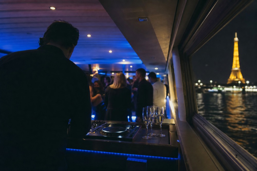 A DJ booth on a Seine dinner cruise with Champagne flutes and a view of the illuminated Eiffel Tower, no text, no logos, no people