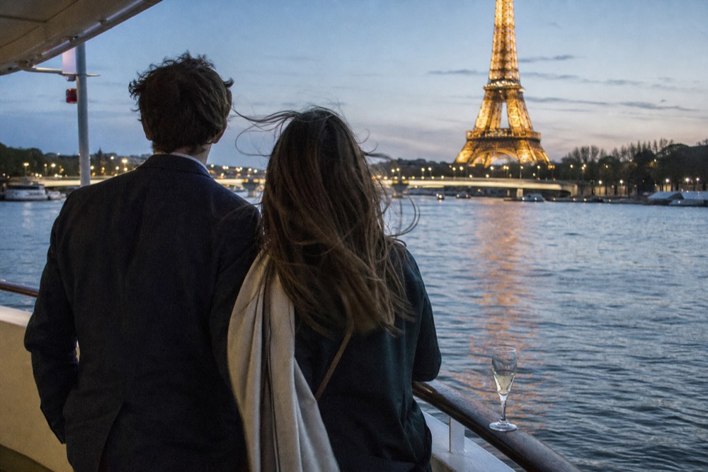 A couple in smart-casual attire on a Seine dinner cruise with the Eiffel Tower in the background, no text, no logos, no people