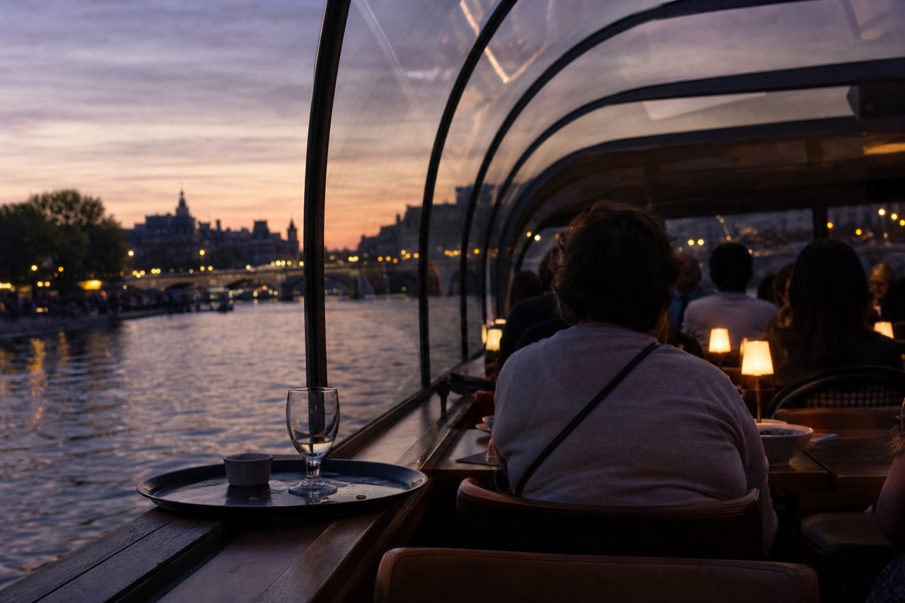 A Seine dinner cruise boat lit at dusk with the Paris skyline and Île de la Cité behind it